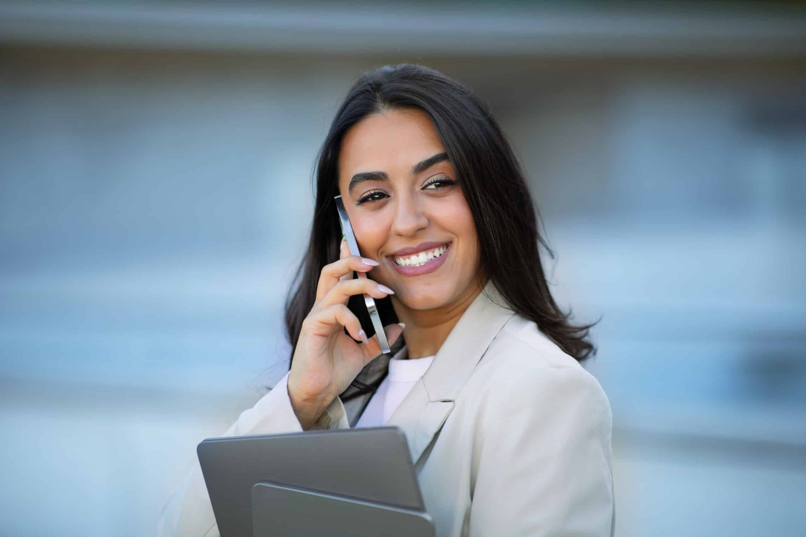 Professional Communication. Portrait of attractive hispanic business lady smiling while communicating on her cellphone outdoors near business center, holding tablet and laptop